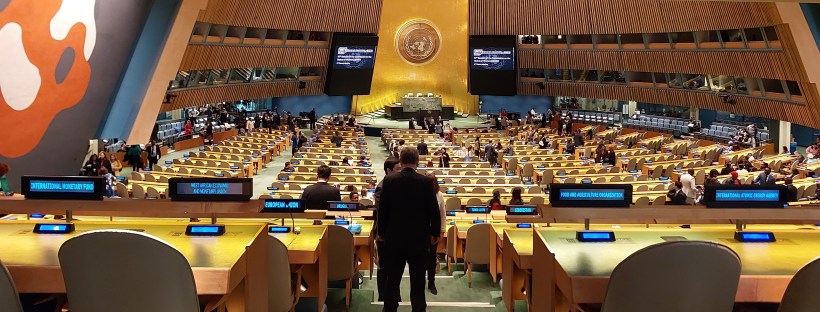 Fotografía del salón principal de asambleas de las Naciones Unidas, el cual es de forma circular. Al fondo, al centro y en lo alto está el logo de la ONU hecho en un metal, de color cobre; está iluminado por una luz blanca que contrasta con el fondo metálico. Además, en lo alto y por los costados hay una serie de ventanas, que es donde están los traductores y traductoras. Bajo el logo, hay un mesón principal levemente más alto, rodeado por pantallas, donde suelen sentarse quienes presiden y moderan la asamblea y luego, hay varias corridas con cientos de sillas y mesones de manera. Cada mesón tiene un letrero digital con el nombre de un país u organización al cual se ha asignado el asiento, además de un micrófono y sistema de sonido. En la sala se ven a algunas personas conversando o de pie durante un receso.